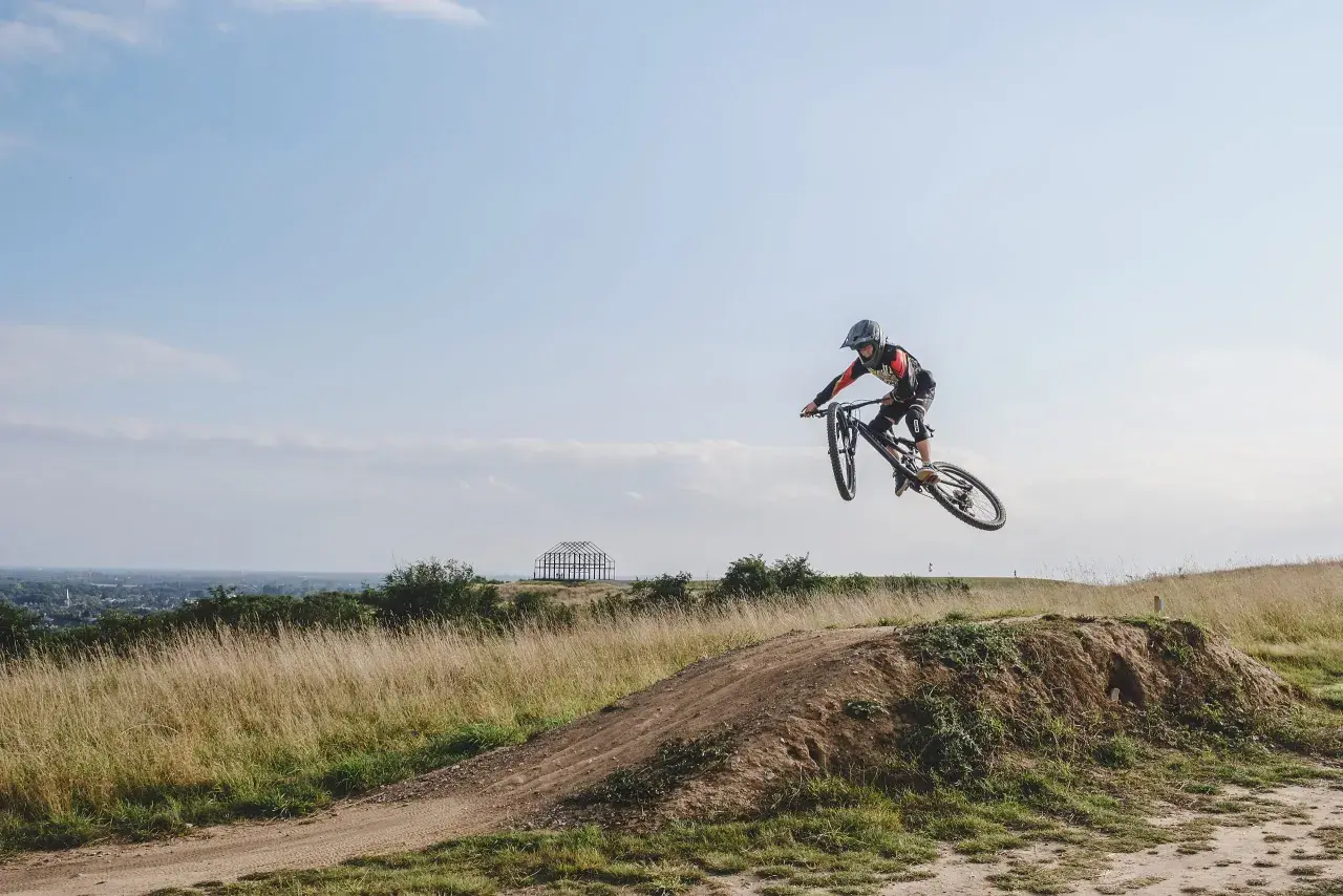 Mountainbiker springt über eine Schanze auf einer mountainbike strecke. Im Hintergrund eine Stadt und eine futuristische Skulptur.