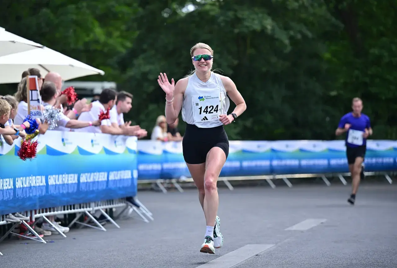 Läuferin mit Startnummer 1424 winkt fröhlich beim Berliner Teamstaffel-Lauf. Zuschauer feuern sie an.