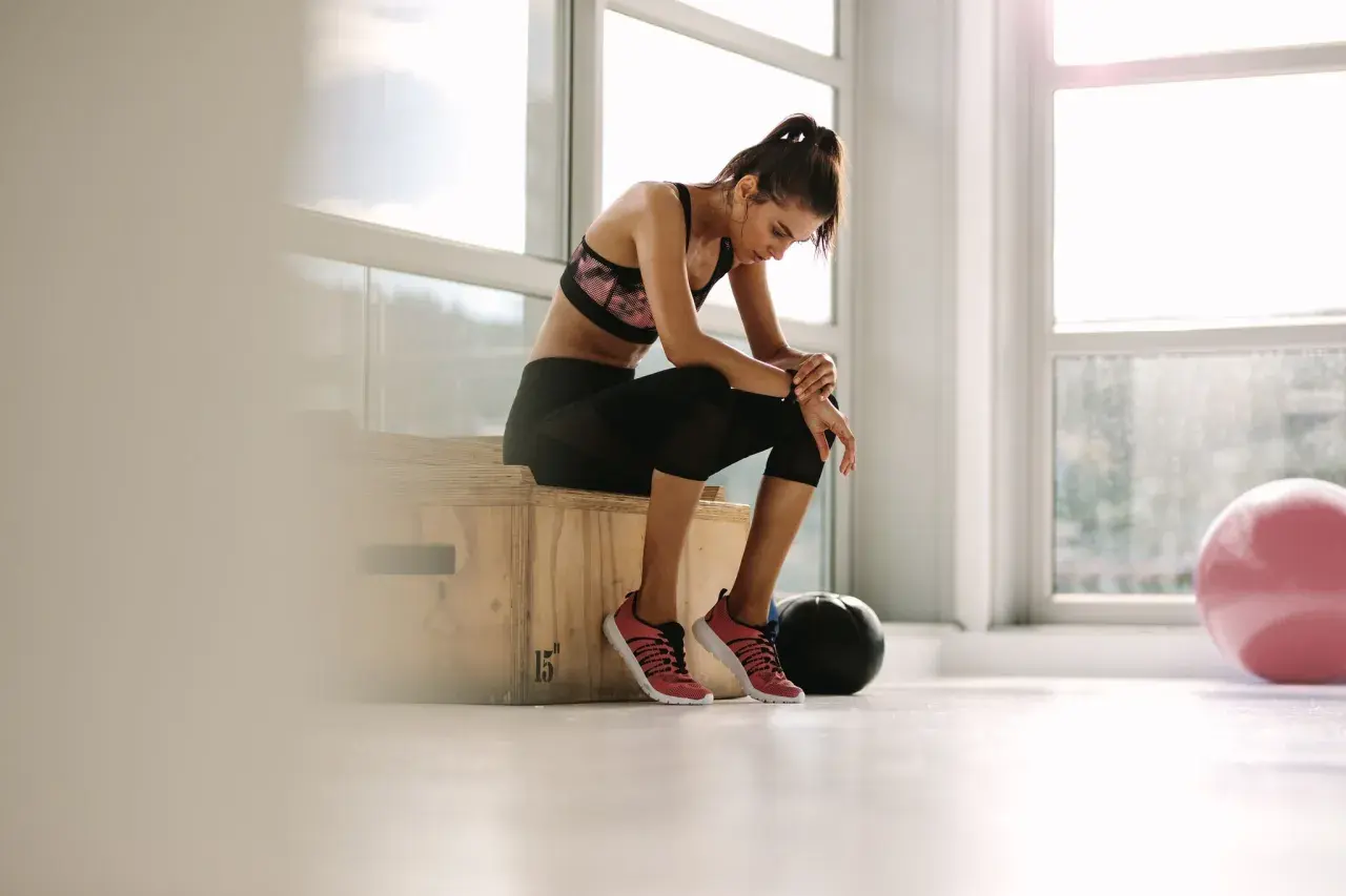 Erschöpfte Frau im **deload gym** sitzt auf einer Box, die Beine angewinkelt, der Kopf gesenkt. Ein schwarzer Medizinball und ein rosa Gymnastikball liegen daneben.