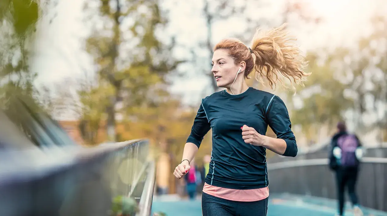 Frau mit Pferdeschwanz, die mit Kopfhörern joggen anfängt. Sie läuft auf einer Brücke, im Hintergrund Bäume und eine andere Person.