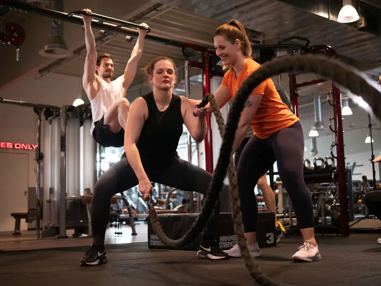 Frauen und Männer trainieren im Fitnessstudio. Eine Frau macht Seilübungen, um richtig zu trainieren. Ein Mann hängt an einer Klimmzugstange.