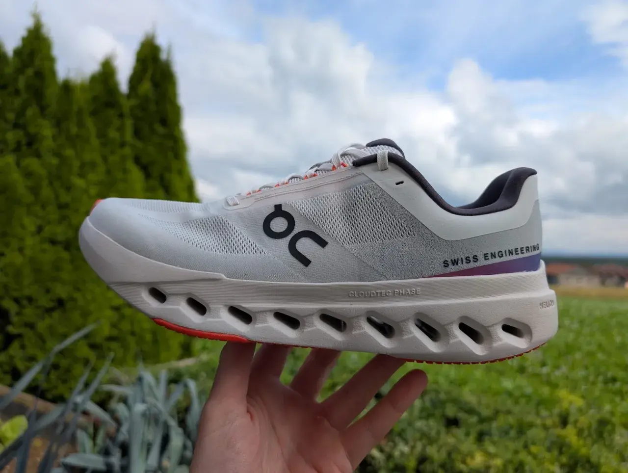 Ein weißer Cloudsurfer next Laufschuh mit "SWISS ENGINEERING" Schriftzug, gehalten in einer Hand vor grüner Natur und blauem Himmel.