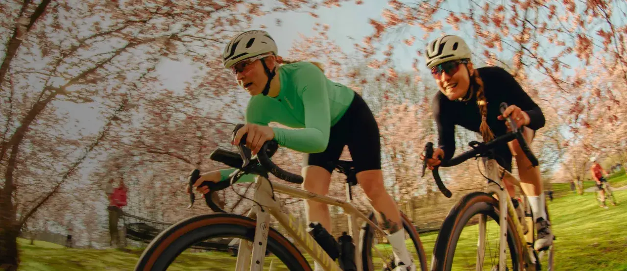 Zwei Frauen auf Rennrädern fahren durch eine blühende Kirschbaumallee. Sie sind Teil einer lebendigen Fahrrad-Community, die die Natur genießt.