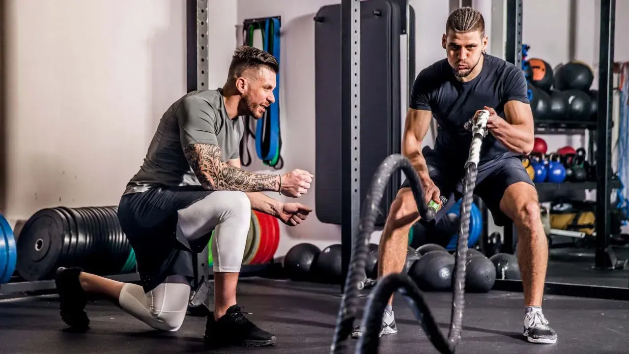 Ein Trainer kniet und erklärt einem Mann, wie er richtig trainieren im Fitnessstudio kann, während dieser mit Battle Ropes arbeitet.