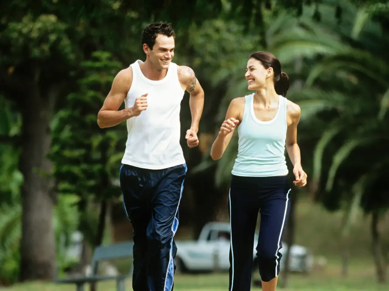 Ein Paar genießt einen Lauf im Park bei angenehmem jogging pace. Sie lachen und schauen sich an, während sie durch das Grün laufen.