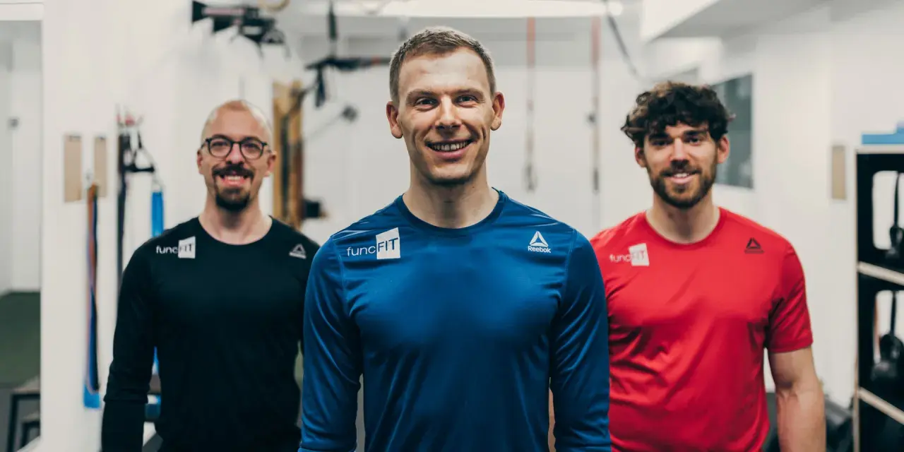 Drei lächelnde Männer in Sportkleidung stehen im Fitnessstudio. Der mittlere Mann, ein fitness trainer, trägt ein blaues Shirt.