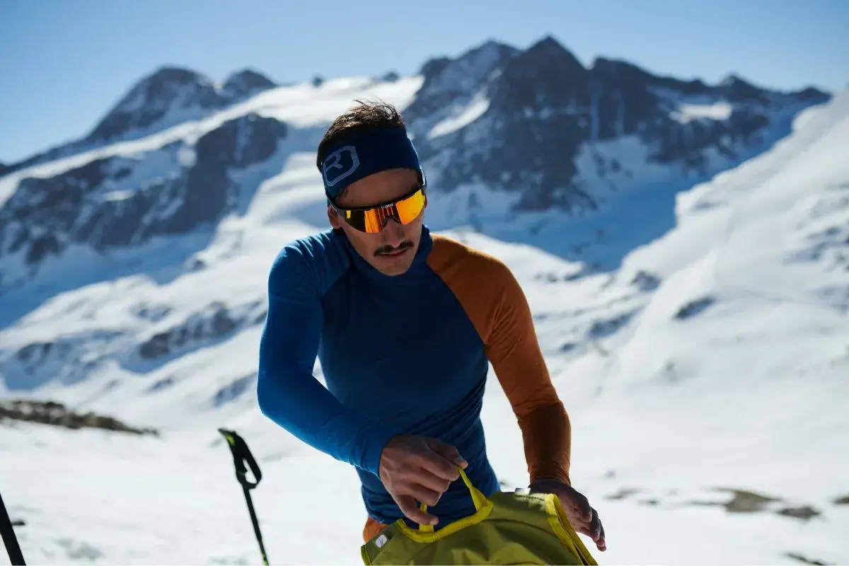 Mann in den Bergen, der sich auf das Zwiebelprinzip Kleidung vorbereitet. Er trägt eine blaue und orangefarbene Funktionsjacke und eine Skibrille.