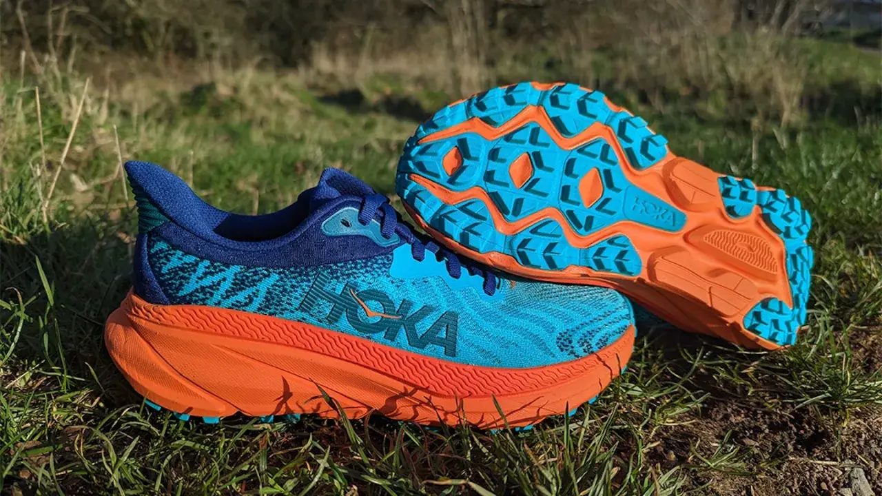 Hoka Challenger 7 Test: Blaue und orangefarbene Trailrunning-Schuhe liegen im Gras. Die Sohle zeigt ein aggressives Profil für Grip.