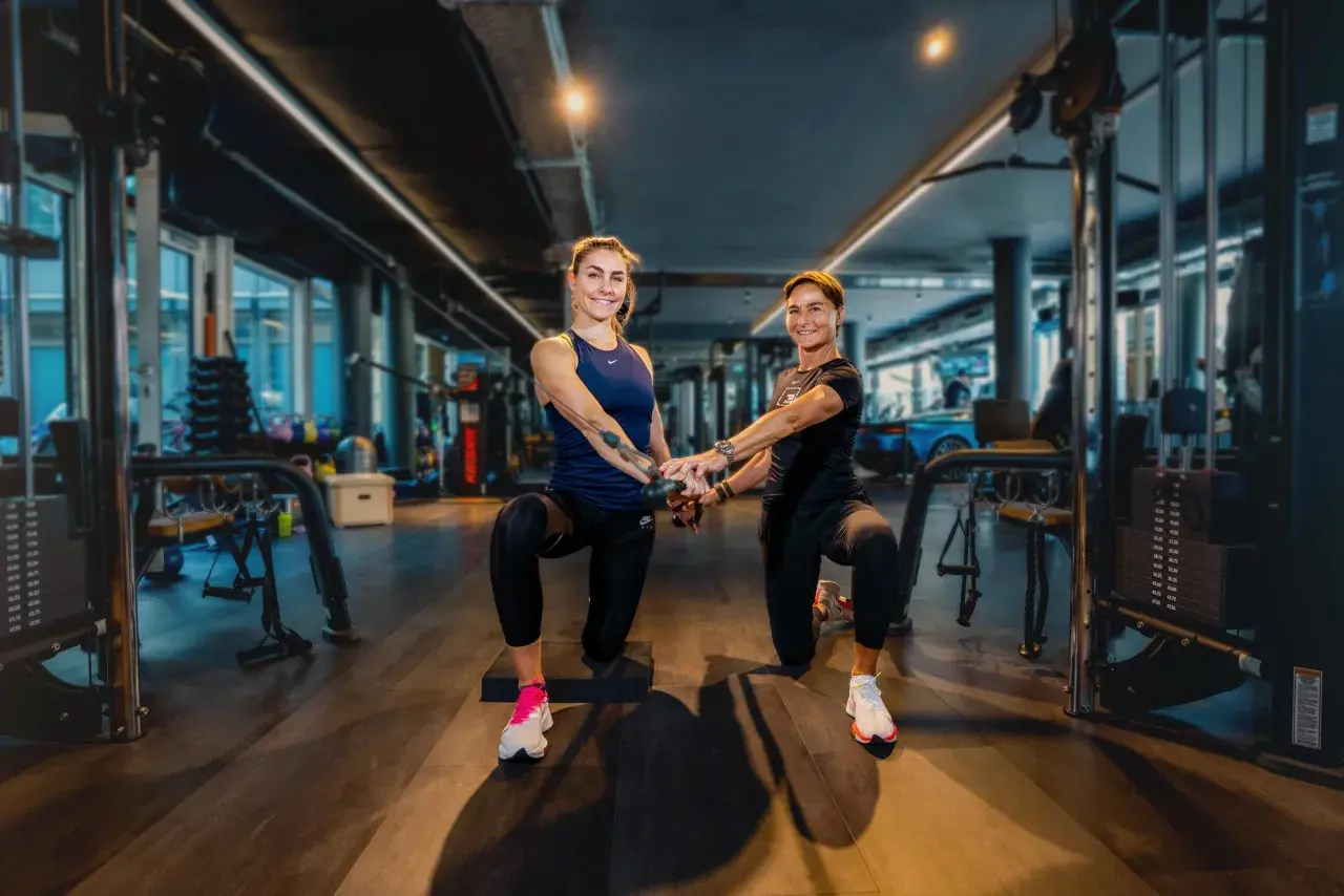 Zwei Frauen trainieren im freihantel bereich eines modernen Fitnessstudios. Sie machen eine Kniebeuge-Übung mit Kabelzug.