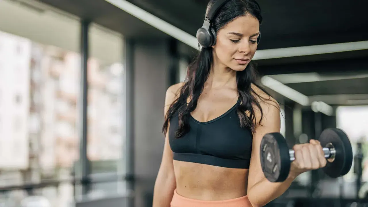 Frau mit Kopfhörern hebt Hantel im Fitnessstudio. Sie fragt sich, wie oft Cardio in der Woche gut ist.