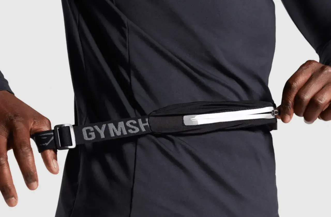 Person schließt eine Bauchtasche, ideal für das handy beim joggen. Der Gurt mit "GYMSH" Schriftzug ist verstellbar.