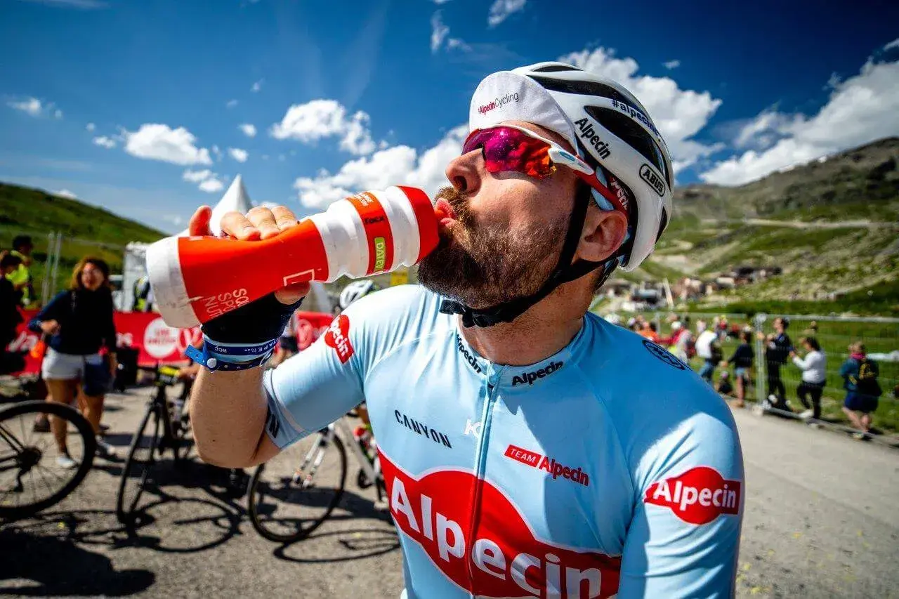 Radfahrer trinkt aus einer Flasche während eines Wettkampfs im Radsport. Er trägt ein hellblaues Trikot mit roten Logos.