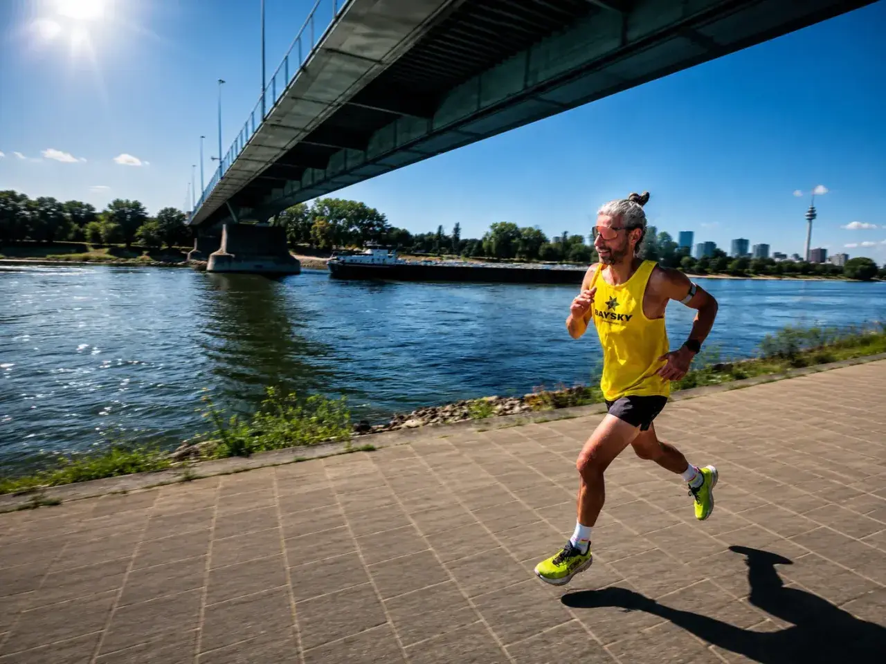 Läufer mit hochrotem Kopf und erhöhtem **Puls bei Hitze** joggt unter einer Brücke am Fluss entlang.