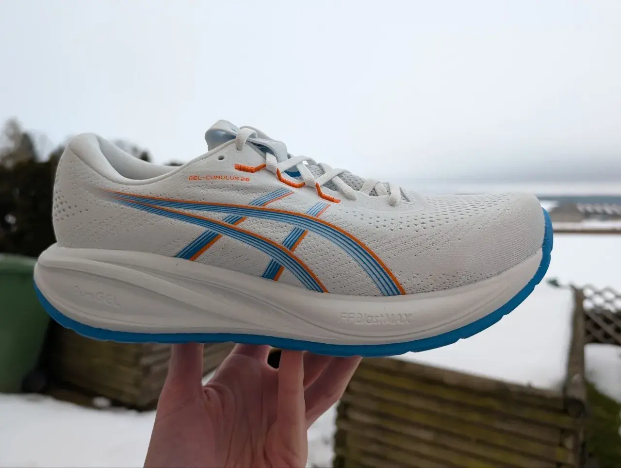 Weißer ASICS GEL-CUMULUS 26 Laufschuh mit blauen und orangen Akzenten, gehalten in einer Hand vor winterlicher Kulisse.