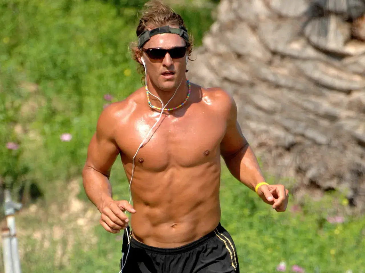 Ein Mann mit Sonnenbrille und Kopfband joggt nackt im Freien. Seine Muskeln glänzen in der Sonne, während er mit Kopfhörern Musik hört.