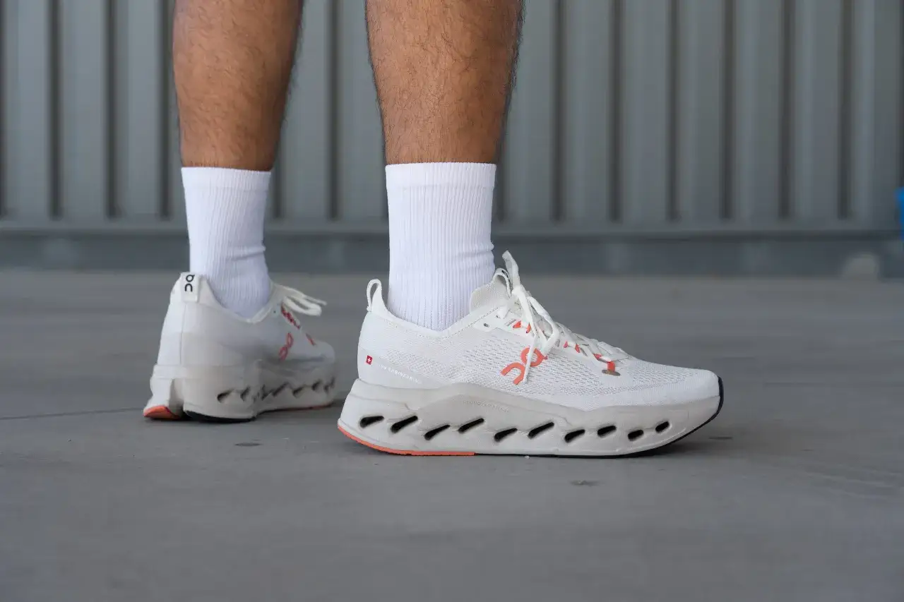 Wei&szlig;e Laufschuhe mit orangefarbenen Akzenten, die auf dem Boden stehen. Die Schuhe sind das Modell Cloudsurfer Max.
