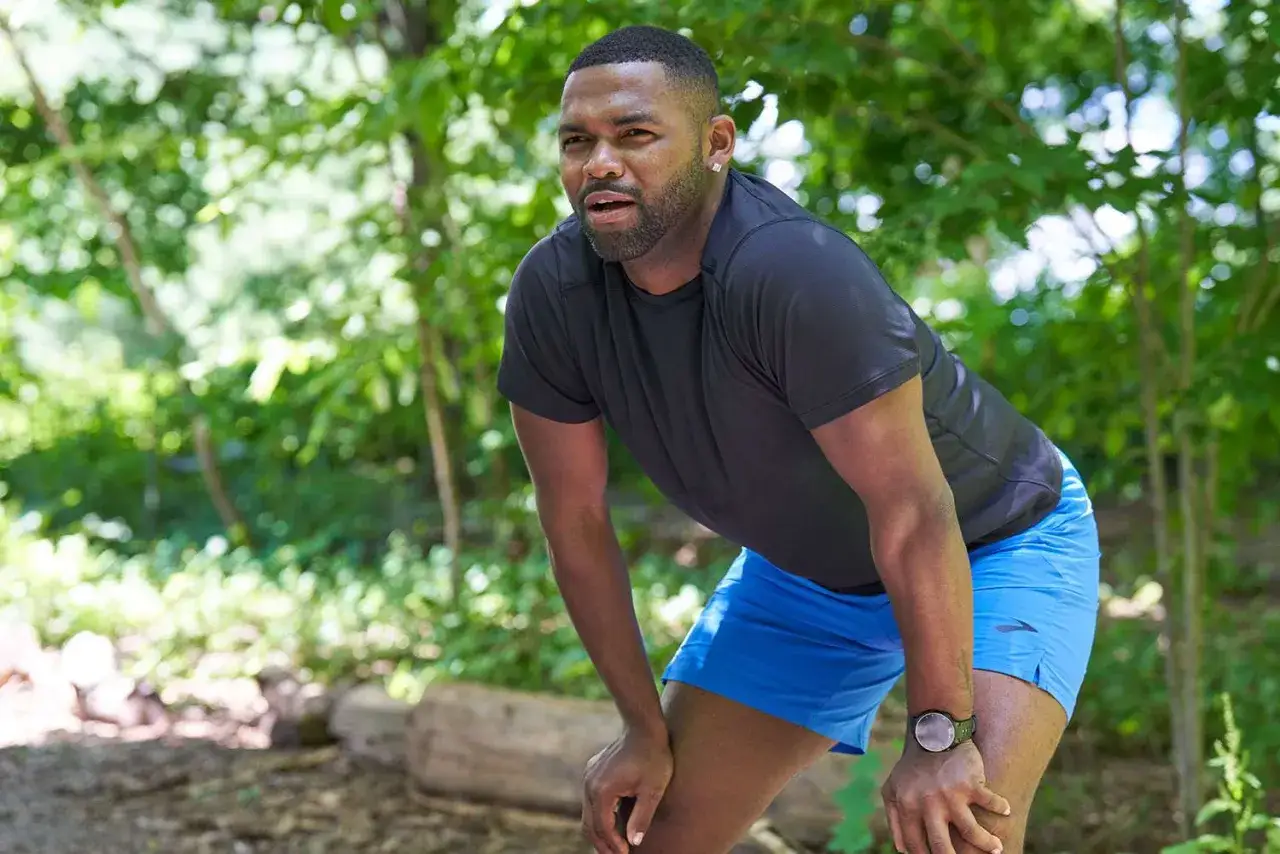 Ein Mann mit Bart und Ohrring im Wald, der sich nach dem Laufen ausruht. Er tr&auml;gt ein schwarzes Shirt und blaue Shorts. Trainingsfreie Tage sind wichtig f&uuml;r die Erholung.