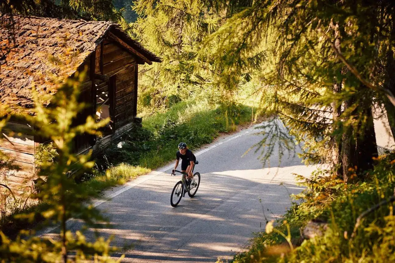 Radfahrer auf einer asphaltierten Stra&szlig;e, vorbei an einer alten Holzh&uuml;tte, inmitten von B&auml;umen. Perfekt f&uuml;r Gravel Bike Touren.