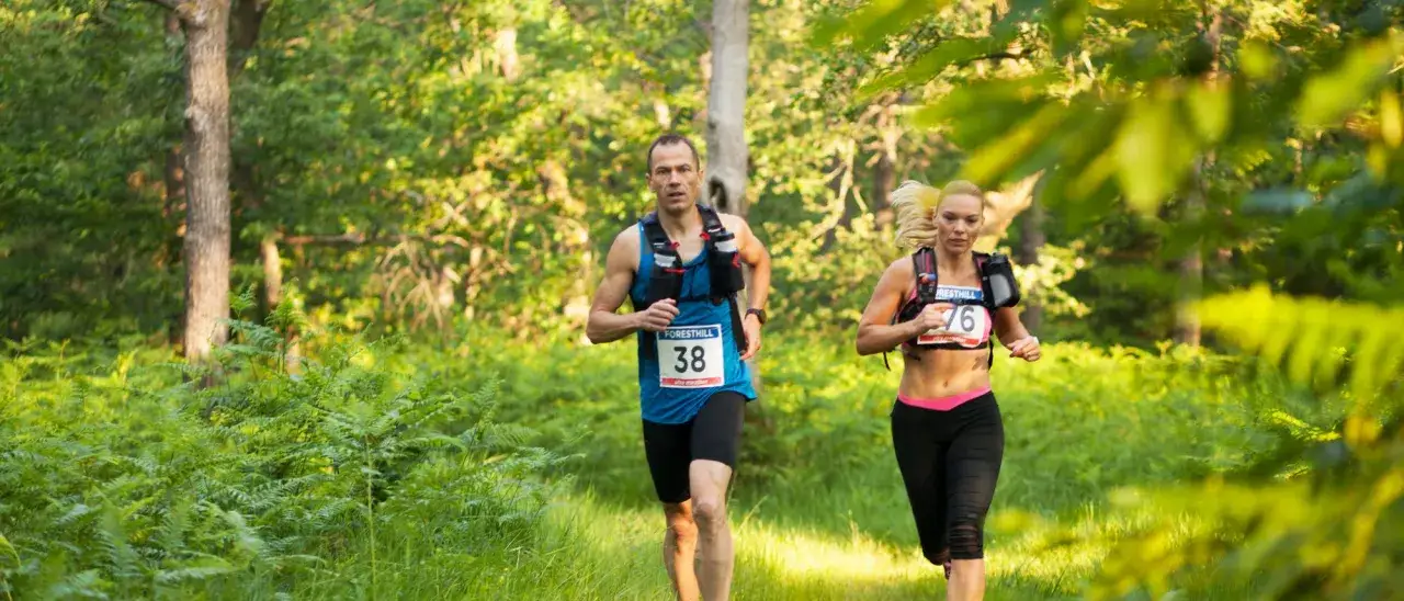 Zwei L&auml;ufer, Mann und Frau, nehmen an einem Ultramarathon Deutschland teil. Sie rennen durch einen sonnendurchfluteten Wald mit Farnen und B&auml;umen.