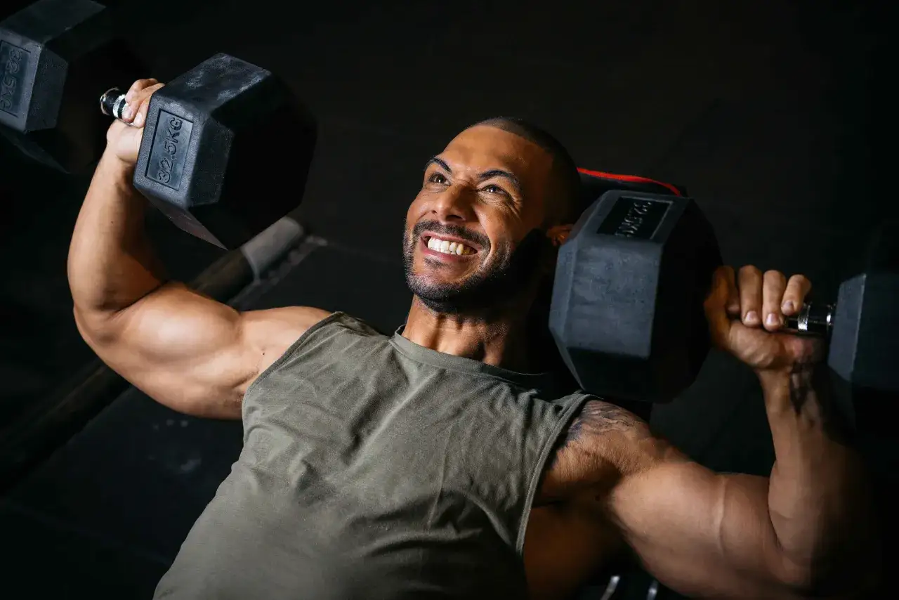 Mann mit kr&auml;ftigen Armen hebt schwere Hanteln (32,5 kg) im SST Training. Sein Gesichtsausdruck zeigt pure Anstrengung und Entschlossenheit.