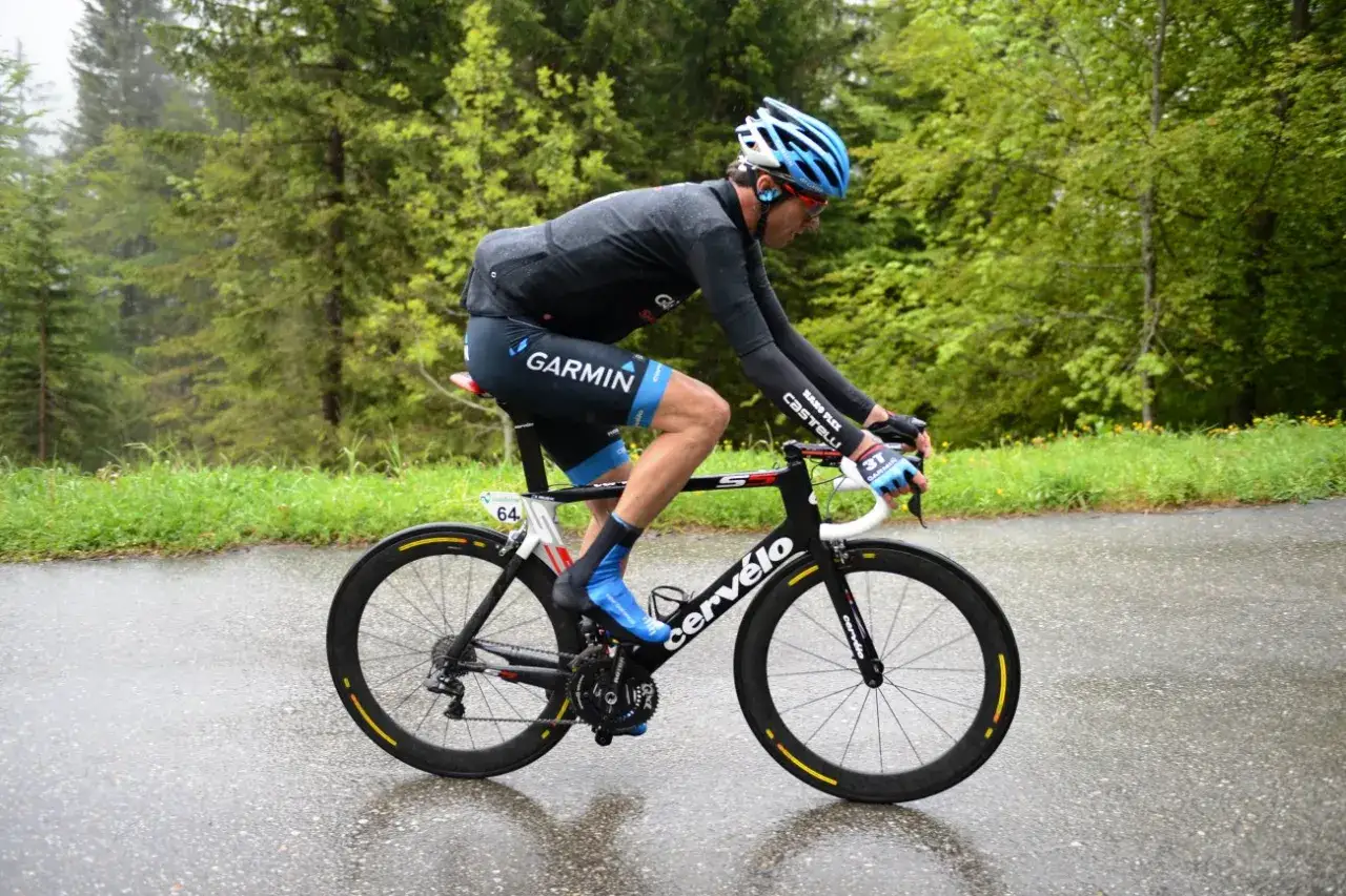 Ein Radfahrer k&auml;mpft sich mit seinem Rennrad bei Regen einen nassen Bergweg hinauf, umgeben von gr&uuml;nen B&auml;umen.