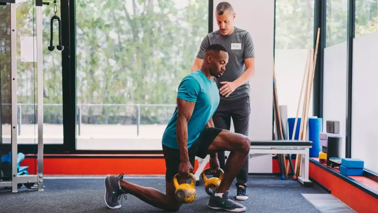 Ein Mann mit Kettlebells macht einen Ausfallschritt, w&auml;hrend ein fitness trainer ihn anleitet.