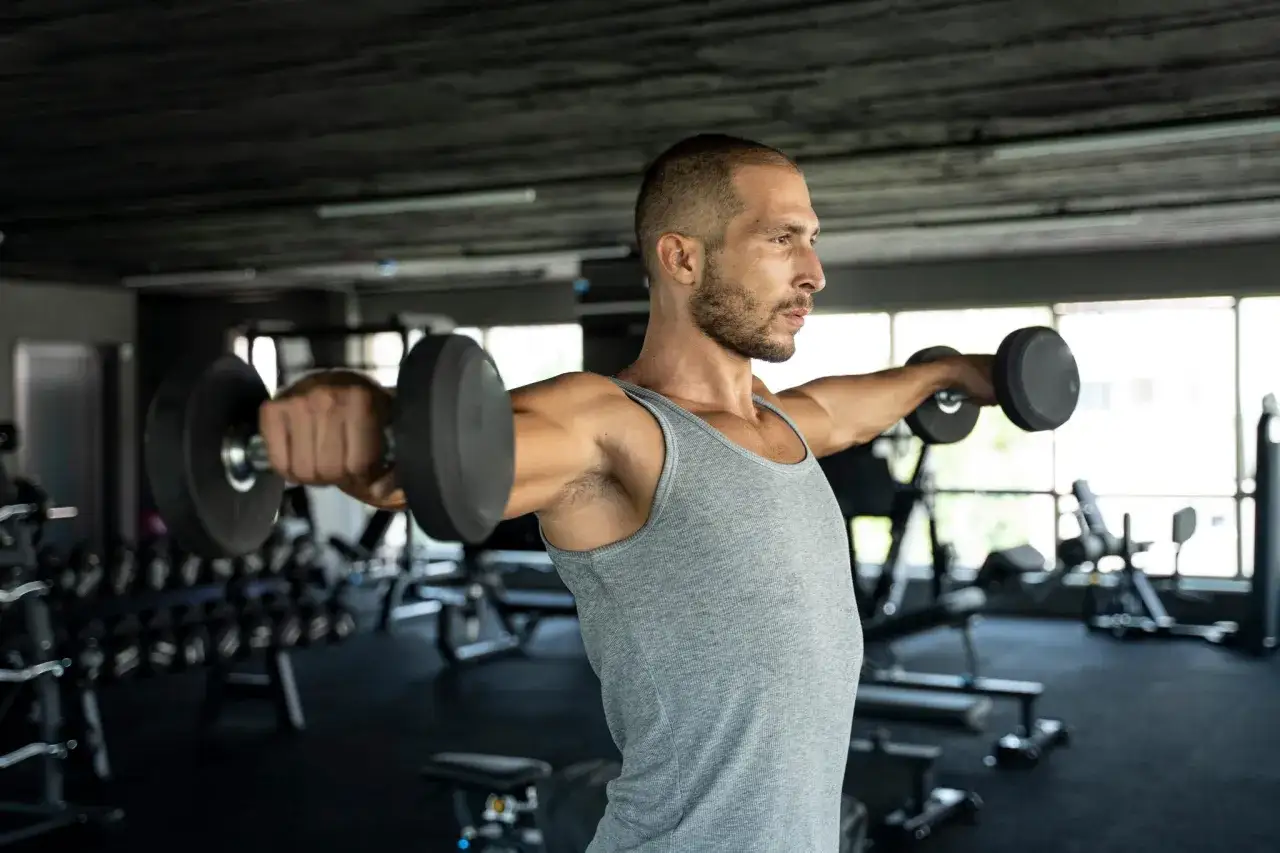 Mann macht Freihantel Training im Fitnessstudio, hebt Gewichte zur Seite.