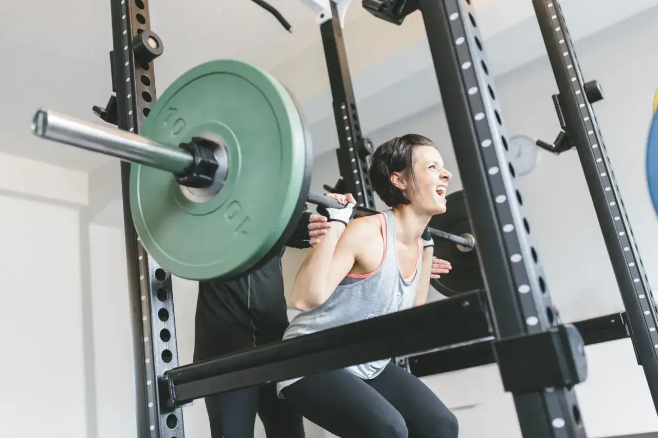 Frau beim Squatten im Fitnessstudio, konzentriert auf ihren Muskelaufbau. Ein Trainer sichert die Hantel.